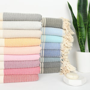 May include: A stack of colorful Turkish towels in various shades, including gray, beige, yellow, pink, and blue. Each towel features a herringbone pattern and fringed edges. A houseplant in a white pot is in the background.