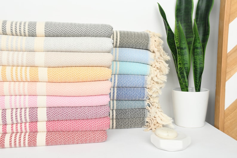 May include: A stack of colorful Turkish towels in various shades, including gray, beige, yellow, pink, and blue. Each towel features a herringbone pattern and fringed edges. A houseplant in a white pot is in the background.