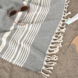 May include: A black and white herringbone patterned beach towel with white striped accents and fringe. Sunglasses, a tube of sunscreen, and a straw sun hat are on the sand next to the towel.