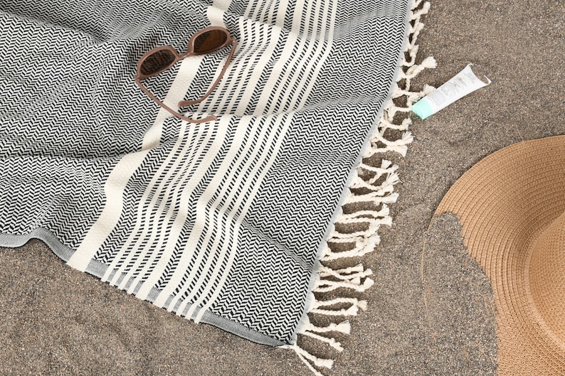 May include: A black and white herringbone patterned beach towel with white striped accents and fringe. Sunglasses, a tube of sunscreen, and a straw sun hat are on the sand next to the towel.