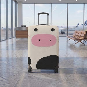 May include: A rolling suitcase featuring a cow-inspired design with a cream-coloured background, black spots, and a large pink oval snout. The luggage has a retractable black handle, four wheels, and a textured surface finish on the exterior shell.
