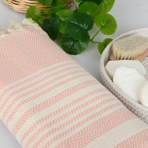 May include: A pink and white striped Turkish towel with fringed edges. A woven basket holds a wooden-handled brush and several white soaps. Green leaves are in the background, suggesting a spa or bathroom setting.