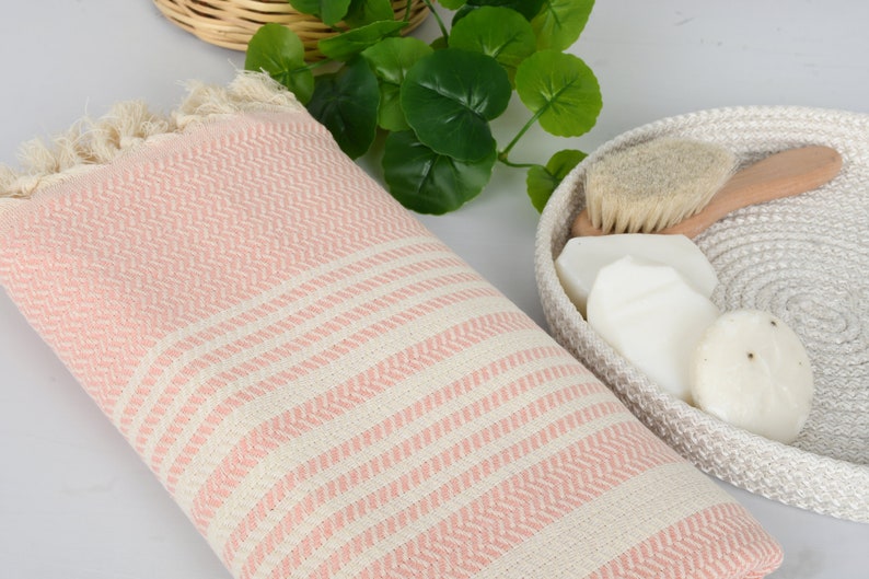 May include: A pink and white striped Turkish towel with fringed edges. A woven basket holds a wooden-handled brush and several white soaps. Green leaves are in the background, suggesting a spa or bathroom setting.