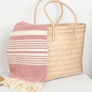 May include: A natural woven tote bag with woven handles, paired with a red and white striped beach towel. The towel has a fringed edge. The bag and towel are on a white surface.