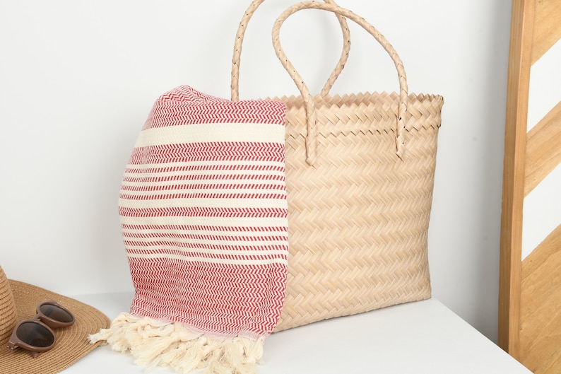 May include: A natural woven tote bag with woven handles, paired with a red and white striped beach towel. The towel has a fringed edge. The bag and towel are on a white surface.