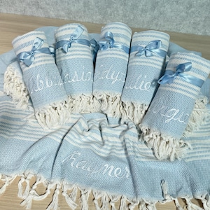 May include: A collection of light blue and white striped Turkish towels, some rolled and tied with light blue ribbons. The rolled towels have names embroidered on them. The flat towel has the name "Rayner" embroidered on it.
