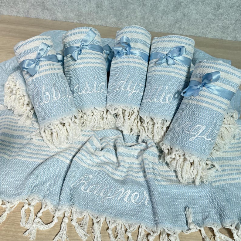 May include: A collection of light blue and white striped Turkish towels, some rolled and tied with light blue ribbons. The rolled towels have names embroidered on them. The flat towel has the name "Rayner" embroidered on it.