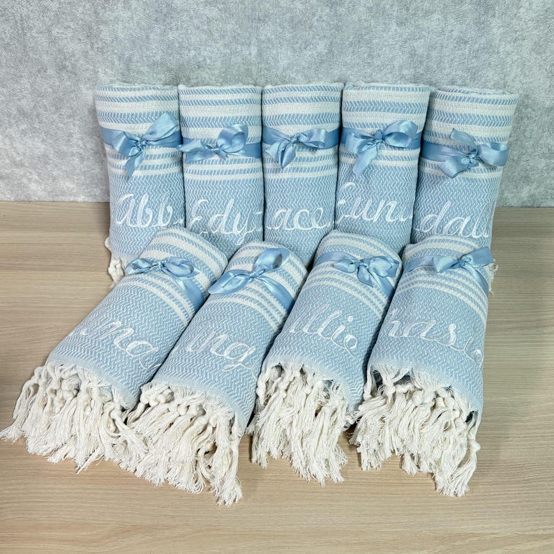 May include: A collection of light blue and white striped towels with white fringe. Each towel is rolled and tied with a light blue ribbon. White embroidered names are visible on the towels. The towels are arranged on a light wood surface.