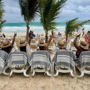 May include: Beach scene with several people in straw hats and gray and white striped towels. The hats have text on them. The towels are draped over white lounge chairs on a sandy beach with palm trees and ocean in the background.
