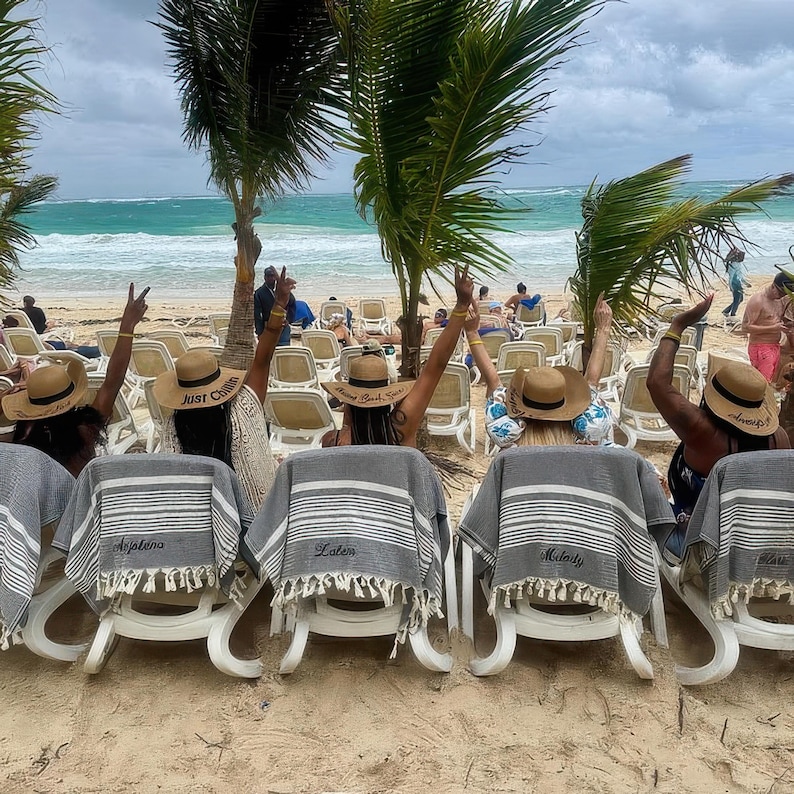 May include: Beach scene with several people in straw hats and gray and white striped towels. The hats have text on them. The towels are draped over white lounge chairs on a sandy beach with palm trees and ocean in the background.