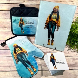 May include: A kitchen set featuring a black oven mitt, a blue and white oven mitt, a blue and white glass cutting board, and a stack of white coasters. The oven mitts and coasters feature a woman wearing a yellow hoodie with the text "Faith Favor Fearless" and a black beanie. The cutting board features the same woman and the text "Faith Favor Fearless". The coasters feature the text "Psalm 90:17 May the favor of the Lord our God rest on us".