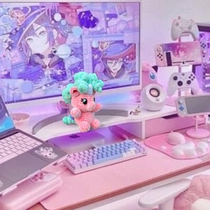 May include: A handmade pink crochet pony with a teal mane and tail. The pony has black eyes and is standing on a pink desk. A computer monitor and gaming accessories are in the background.