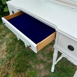 Pode incluir: Uma penteadeira branca com uma gaveta aberta, revelando um interior azul marinho. A gaveta &eacute; de madeira e tem uma ma&ccedil;aneta de lat&atilde;o. A mesa tem um espelho e est&aacute; sobre a grama.