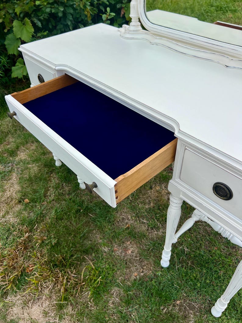 Pode incluir: Uma penteadeira branca com uma gaveta aberta, revelando um interior azul marinho. A gaveta &eacute; de madeira e tem uma ma&ccedil;aneta de lat&atilde;o. A mesa tem um espelho e est&aacute; sobre a grama.