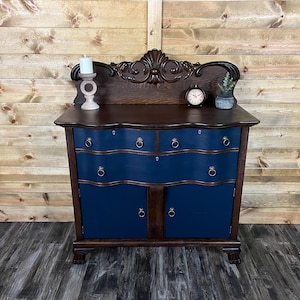 May include: A dark wood antique dresser with a navy blue front. The dresser has four drawers and two doors with brass ring pulls. A decorative wooden accent sits atop the dresser, along with a small clock, candle holder, and a small vase.
