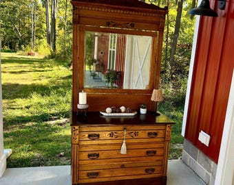 May include: A vintage wooden dresser with a large mirror, featuring ornate carvings and three drawers. The dresser is a warm brown color, with a dark top and a decorative tassel. A small lamp and candle sit on top.