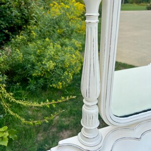 Pode incluir: Um espelho de toucador branco com um acabamento envelhecido. O espelho tem uma base e uma moldura decorativas e ornamentadas com uma borda prateada. O espelho &eacute; colocado em um fundo de folhagem verde e grama.