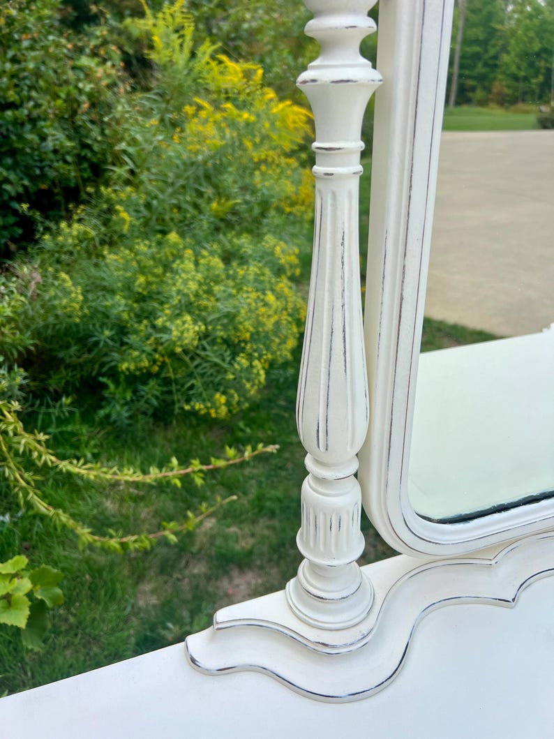 Pode incluir: Um espelho de toucador branco com um acabamento envelhecido. O espelho tem uma base e uma moldura decorativas e ornamentadas com uma borda prateada. O espelho &eacute; colocado em um fundo de folhagem verde e grama.