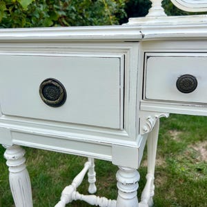 Pode incluir: Toucador branco sujo com duas gavetas e detalhes ornamentados. As gavetas t&ecirc;m puxadores de cor bronze escuro. A mesa tem um acabamento envelhecido, dando-lhe um aspecto vintage ou antigo. As pernas s&atilde;o torneadas e a mesa est&aacute; sobre uma superf&iacute;cie gramada.