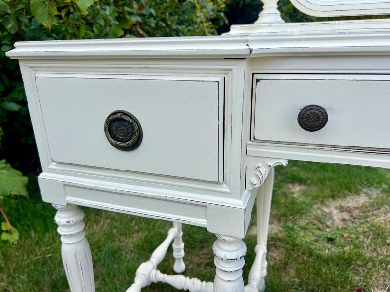 Pode incluir: Toucador branco sujo com duas gavetas e detalhes ornamentados. As gavetas t&ecirc;m puxadores de cor bronze escuro. A mesa tem um acabamento envelhecido, dando-lhe um aspecto vintage ou antigo. As pernas s&atilde;o torneadas e a mesa est&aacute; sobre uma superf&iacute;cie gramada.