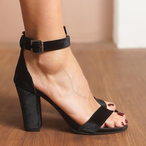 Puede incluir: Sandalia negra de tacón alto de terciopelo con tacón grueso y correa en el tobillo. La sandalia tiene un cierre de hebilla y una sola correa en los dedos. El zapato está sobre un suelo de madera.