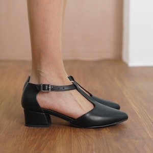Profile view of a model wearing Hyl black vegan leather T-strap mary jane heels, showcasing the pointed toe and low block heel.