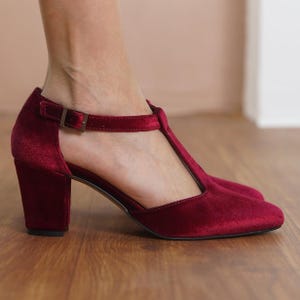 Puede incluir: Zapatos de tacón de terciopelo burdeos con tira en T y cierre de hebilla.