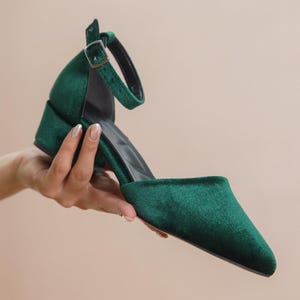 Hand holding a pair of green velvet block-heeled ankle strap shoes, highlighting the sleek design and adjustable buckle.