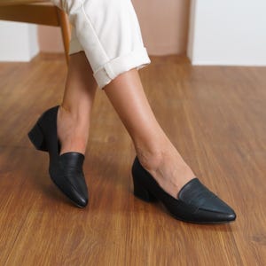 Top down flat lay of two black vegan leather loafers, showcasing the elegant pointed toe design and contrasting sturdy low block heels, arranged on a neutral background.