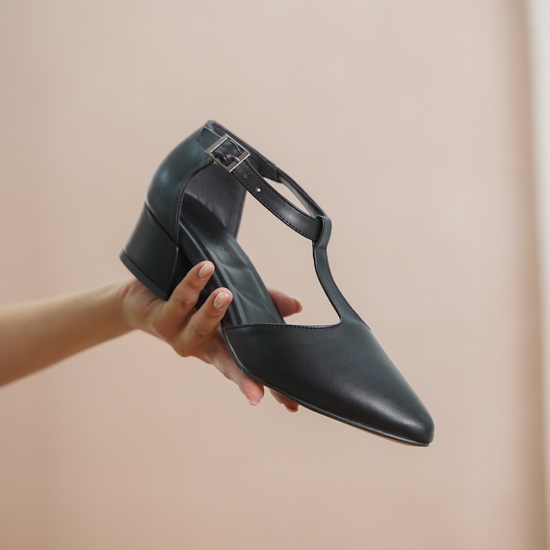 Close-up of hands holding a pair of Hyl black vegan leather T-strap mary jane heels, highlighting the luxurious material and chic design.