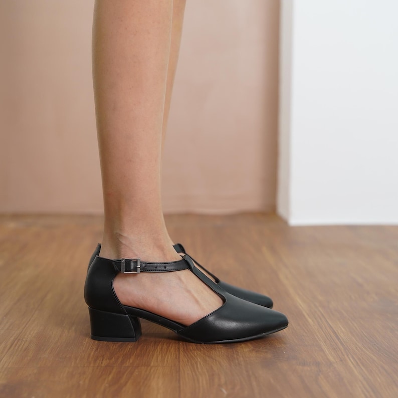 Profile view of a model wearing Hyl black vegan leather T-strap mary jane heels, showcasing the pointed toe and low block heel.