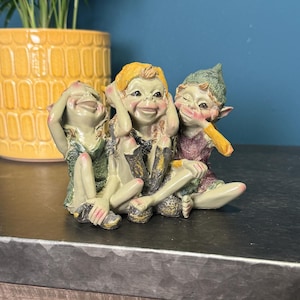 May include: Three small, colourful figurines of mythical creatures, each with a different expression. One covers its eyes, one covers its ears, and one covers its mouth. The figurines are sitting on a dark grey surface.