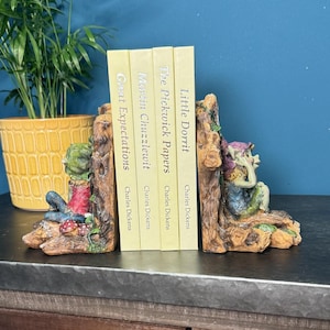 May include: A pair of bookends featuring a whimsical design of two gnomes holding books. The bookends are made of a light brown material and have a rustic finish. The books are titled "Little Dorrit", "The Pickwick Papers", "Martin Chuzzlewit", and "Great Expectations" by Charles Dickens.