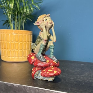 May include: A green and brown figurine of a gnome with large ears, sitting on a red and yellow mushroom with white spots. The gnome is holding its hands to its ears and has a surprised expression on its face.