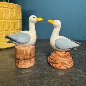 May include: A pair of ceramic seagull salt and pepper shakers. The seagulls are white with blue wings and yellow beaks. They are perched on brown bases, one with a rope and the other with a wooden post.
