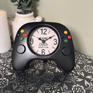 May include: Black gamepad-shaped clock with a white clock face. The clock face has black numbers and hands, with the words "GAME OVER INSERT COIN". Coloured buttons are on the top and sides of the clock.
