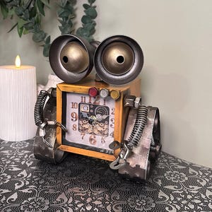 May include: A quirky steampunk robot clock. The clock features a yellow wooden body, metal wheels, and large, round, gold-coloured eyes. The clock face displays Roman numerals. A unique decorative item, the clock measures approximately 15 cm in height.
