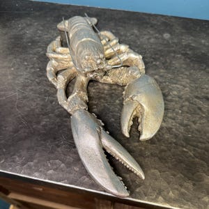 May include: A silver lobster sculpture with detailed claws, body segments, and antennae. The lobster is positioned on a dark, textured surface, showcasing its intricate design and metallic finish. A decorative nautical theme.