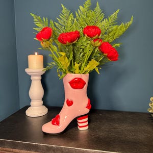 May include: A pink ceramic boot-shaped vase with red lips, hearts, and a striped heel. The vase contains red flowers and green foliage. A white candle holder with a lit candle is to the left.