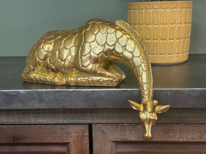 May include: A gold-colored giraffe figurine with a textured, scaly surface. The giraffe is lying down with its head bent down, as if it is looking at something.