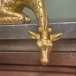 May include: A gold-colored giraffe figurine with a textured surface. The giraffe is in a crouching position with its head bent down.
