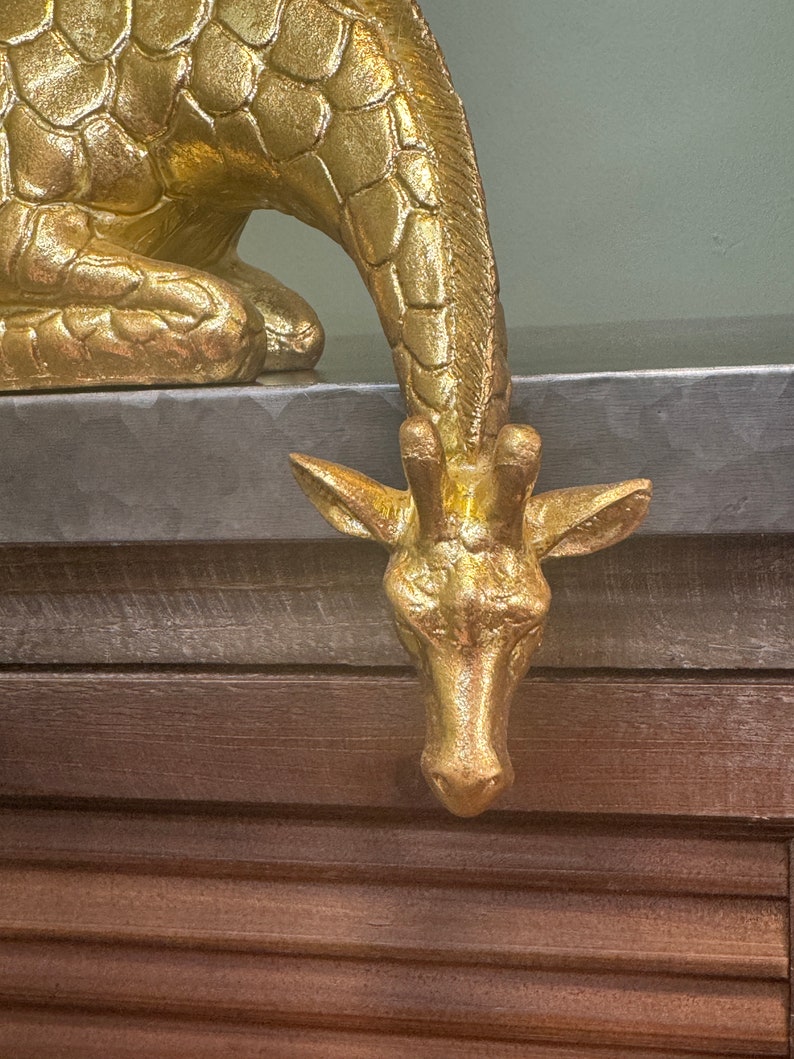 May include: A gold-colored giraffe figurine with a textured surface. The giraffe is in a crouching position with its head bent down.