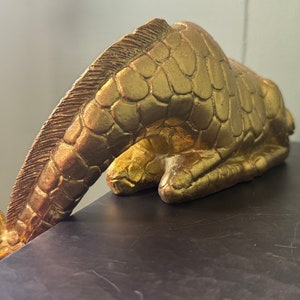 May include: A gold-colored giraffe figurine with a textured surface, lying on its side with its head bent down.