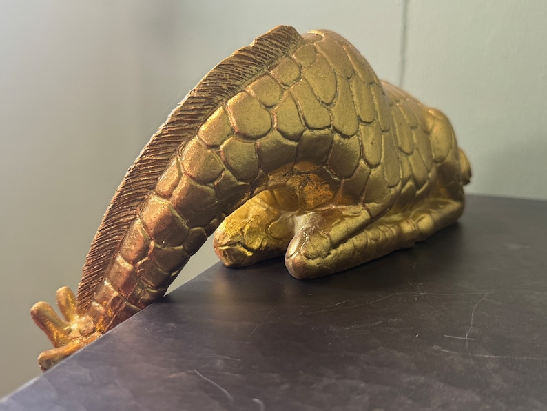 May include: A gold-colored giraffe figurine with a textured surface, lying on its side with its head bent down.
