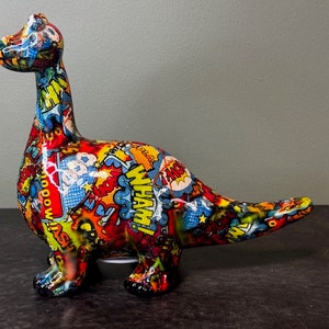 May include: A colourful ceramic dinosaur money box with a comic book design. The dinosaur is red, yellow, blue, and green with white and black accents. The design features words like "POW!" and "BAM!"