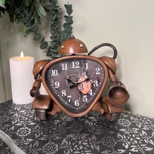 May include: A brown robot-shaped clock with a dark clock face. The clock has a round head, a triangular body, and arm-like structures. The clock face features black hands and white numbers. The clock is placed on a patterned surface.