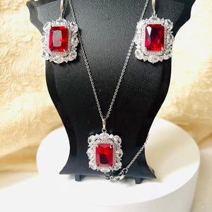 May include: A silver-toned jewellery set comprising a necklace and earrings. Each piece features a large, rectangular, red gemstone framed by smaller, clear stones. The set is displayed on a black stand against a neutral backdrop.