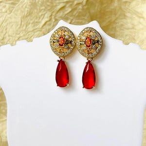 Può includere: Un paio di orecchini color oro con un design circolare, con pietre preziose chiare e arancioni. Ogni orecchino ha un pendente a forma di goccia, in pietra rosso scuro. Gli orecchini sono esposti su un busto bianco.