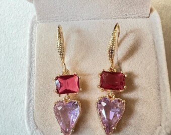 May include: A pair of gold earrings with pink and red heart-shaped crystals. The earrings have a dangling design with a square-shaped crystal above the heart-shaped crystal.