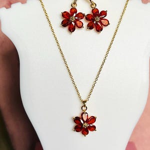May include: A gold necklace and earrings set with red flower-shaped pendants. The necklace has a delicate chain and the earrings are dangle style.